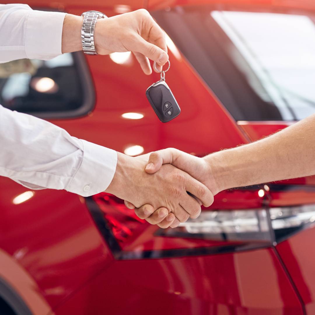 Two people shaking hands while one person hands the other a black and silver key fob in front of a shiny, red car.