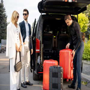 A man and woman wearing business attire watch a chauffeur load their luggage into a vehicle. The chauffeur wears all black.