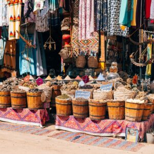 A colorful outdoor market stand with a variety of clothing hanging above a display of baskets, sitting on traditional fabric.