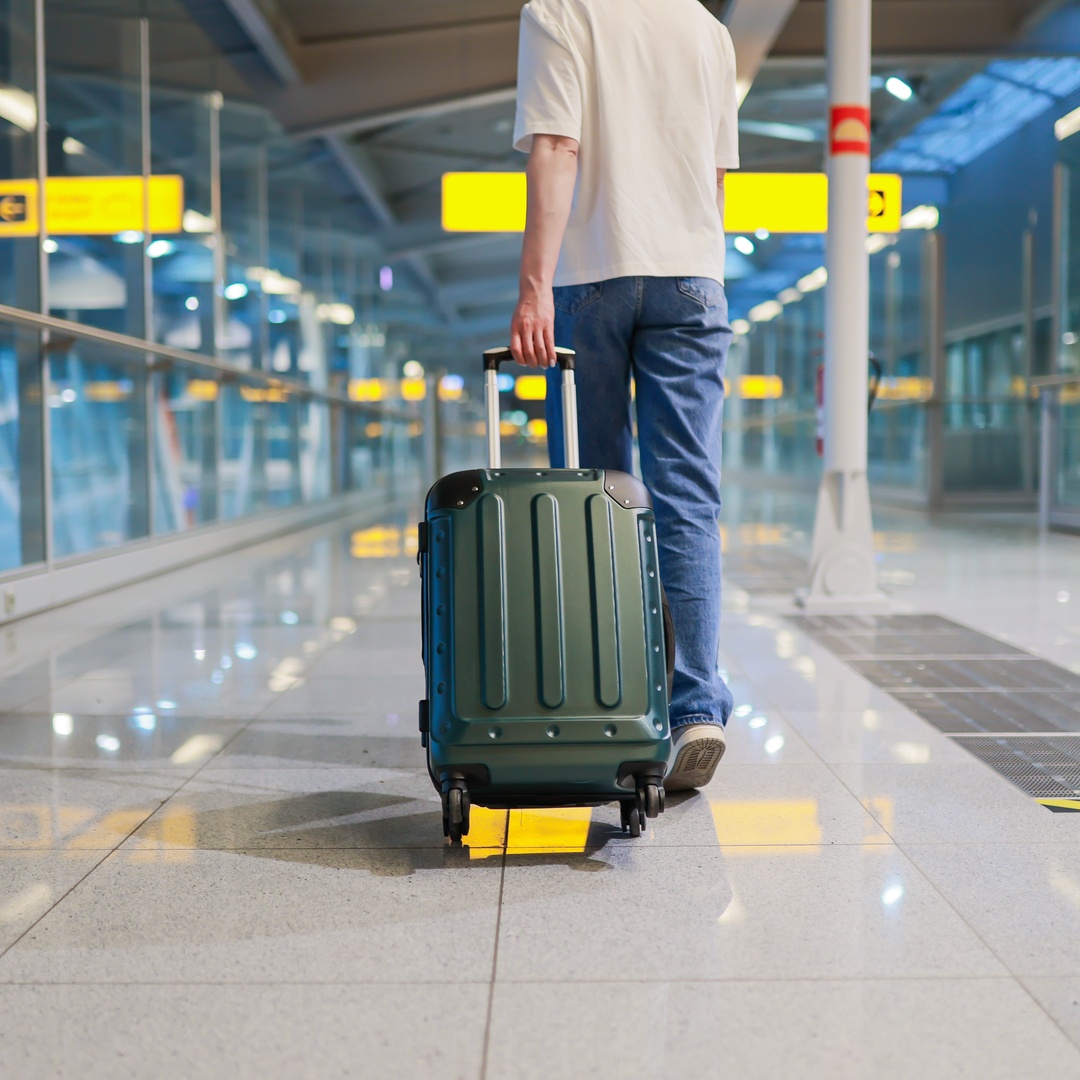 Someone in jeans and a T-shirt rolls a green carry-on through an airport. A yellow sign with an arrow is ahead of them.