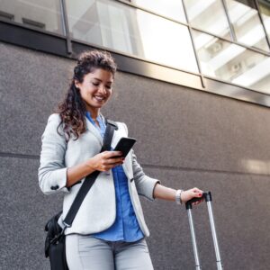 A woman in professional attire with a laptop bag over her shoulder and a hand on a suitcase smiles down at her phone.