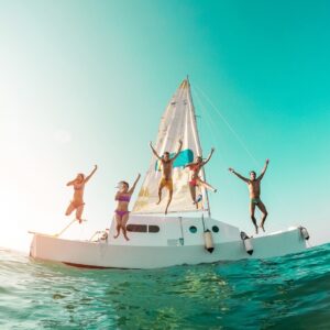 A group of young people in swimsuits leaping from a white sailing catamaran into turquoise water on a sunny day.