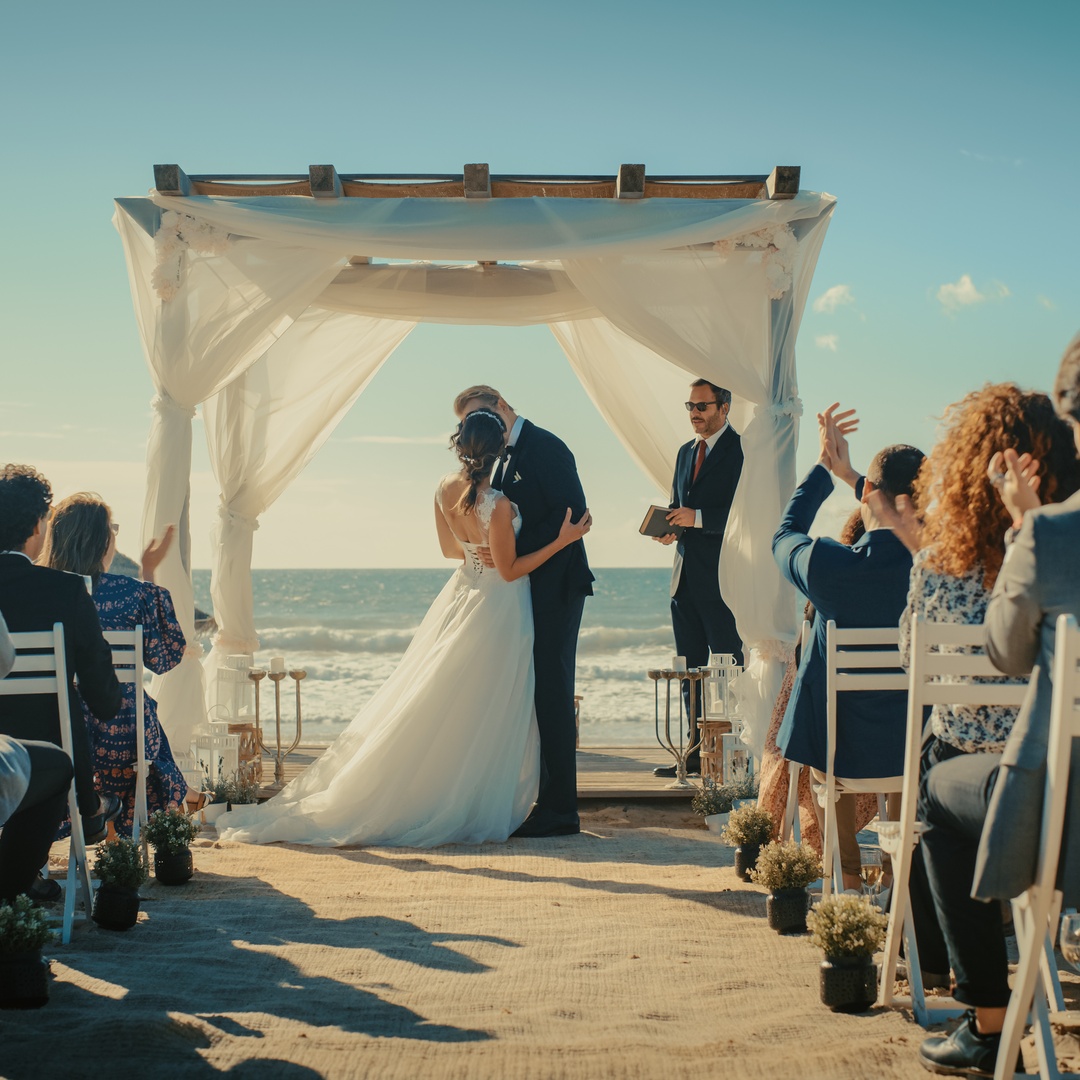A bride and groom kiss at their beach wedding ceremony. The guests clap while looking toward the couple.