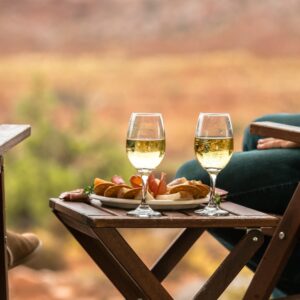 Two people relaxing outdoors on wooden chairs with a small folding table holding wine glasses and charcuterie between them.