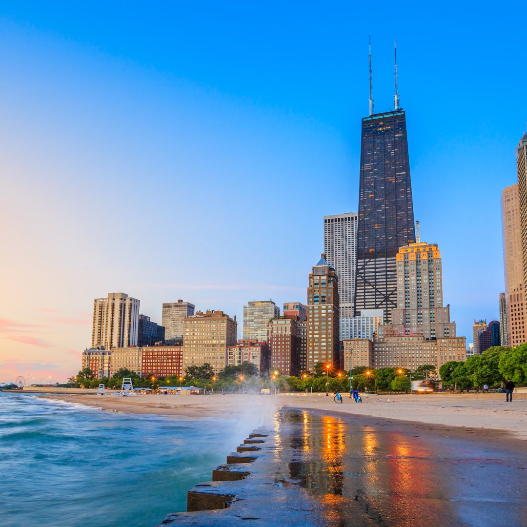 A concrete walkway that meets the beach along Lake Michigan. Behind the beach is part of the Chicago skyline.