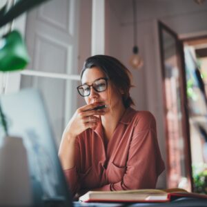 A young businesswoman is thinking about something while sitting in front of an open portable laptop in her office.