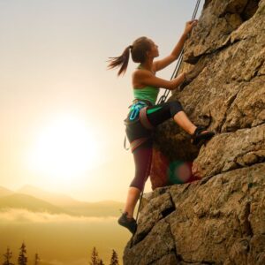 A woman looks up as she climbs a large rock in the mountains, the bright sun low in the sky behind her.