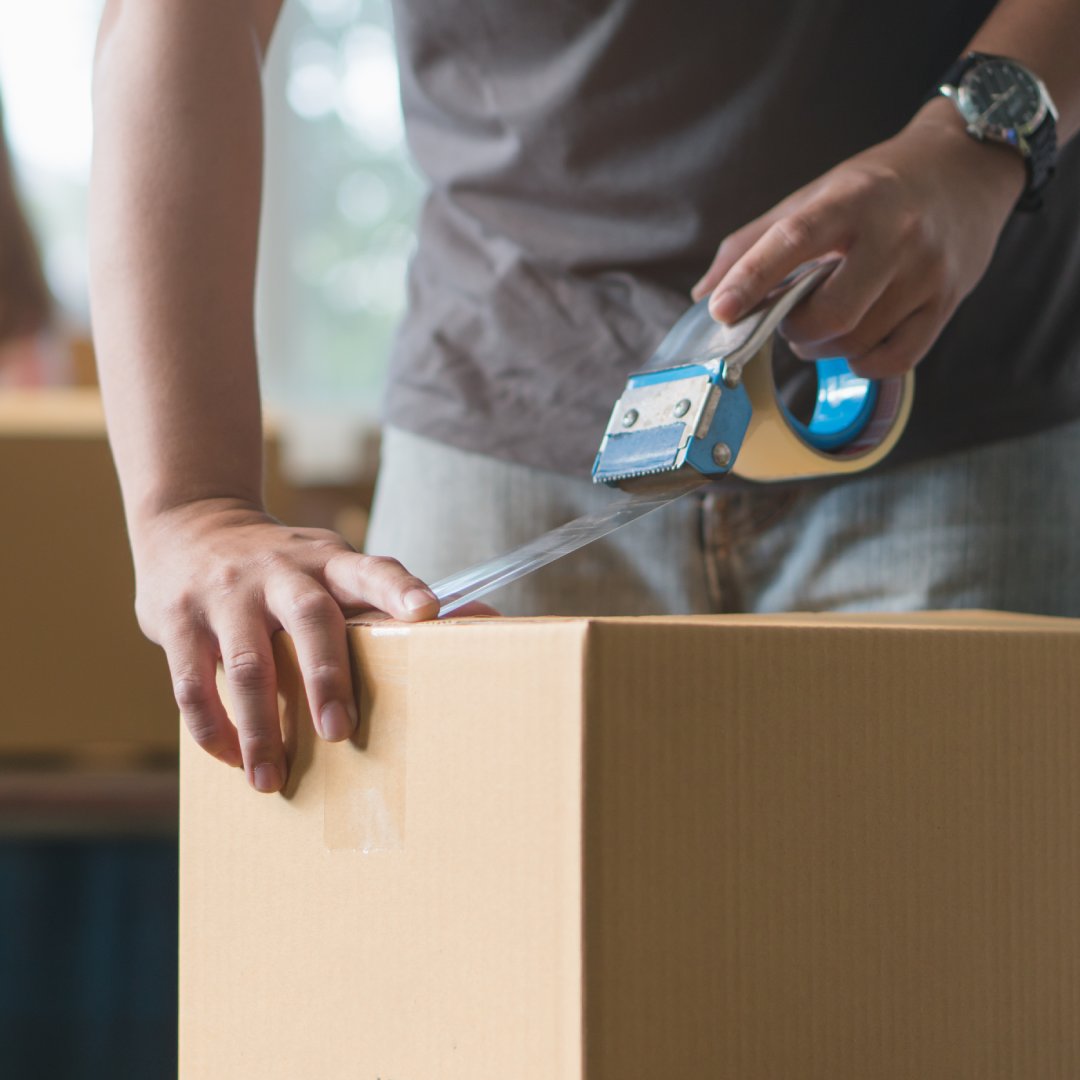A close up of a person sealing a cardboard moving box with a roll of clear packing tape in a house.