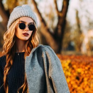 A woman wearing a black shirt, gray jacket, gray beanie, and round black sunglasses stands in front of a fall landscape.