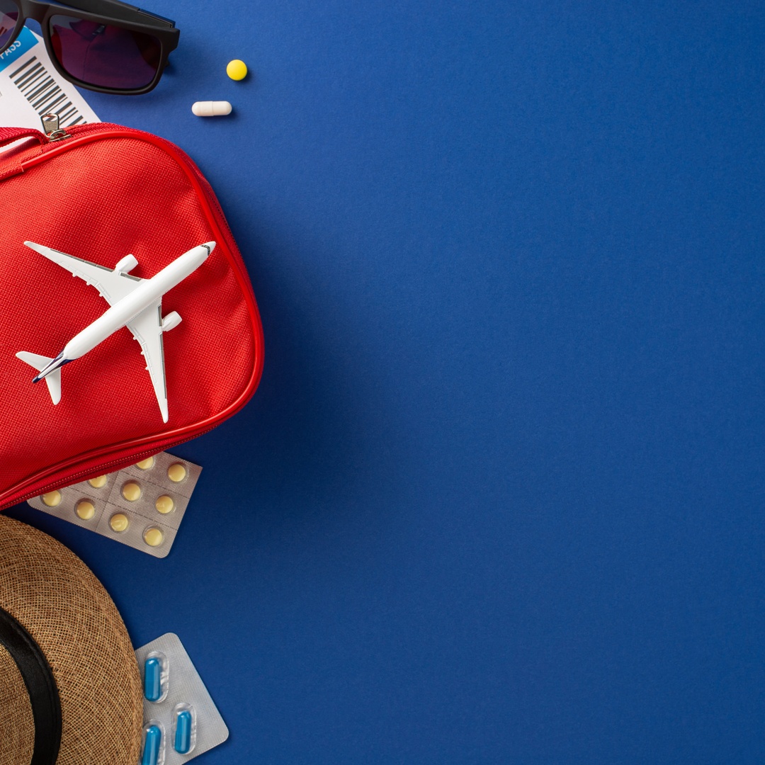 A first aid kit, medication, a boarding pass, a passport, sunglasses, and a hat sit on a blue background.
