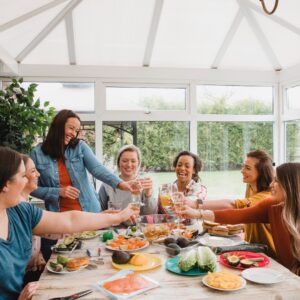 Several women are seated around a table laden with plates of food. They are raising their glasses in a toast.