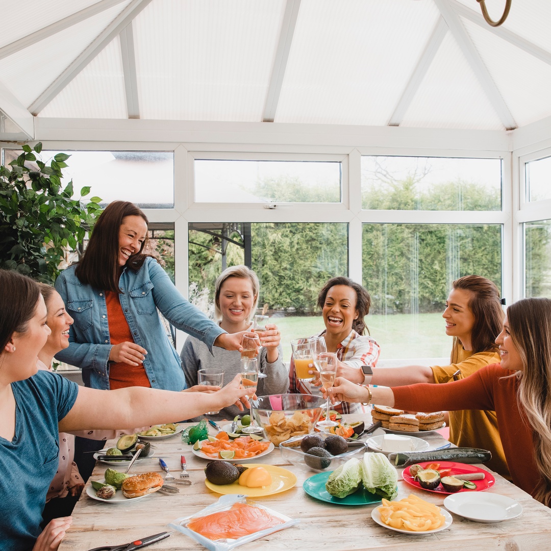 Several women are seated around a table laden with plates of food. They are raising their glasses in a toast.