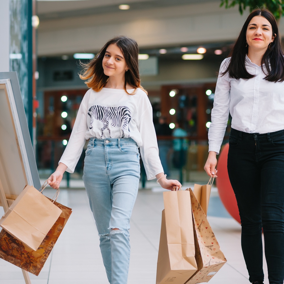 A teenage girl and her mother are smiling and laughing as they shop at the mall, holding many shopping bags.