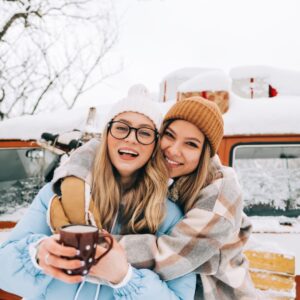 Two women smiling in front of a snow-covered van. They are both wearing winter clothes to keep themselves warm.
