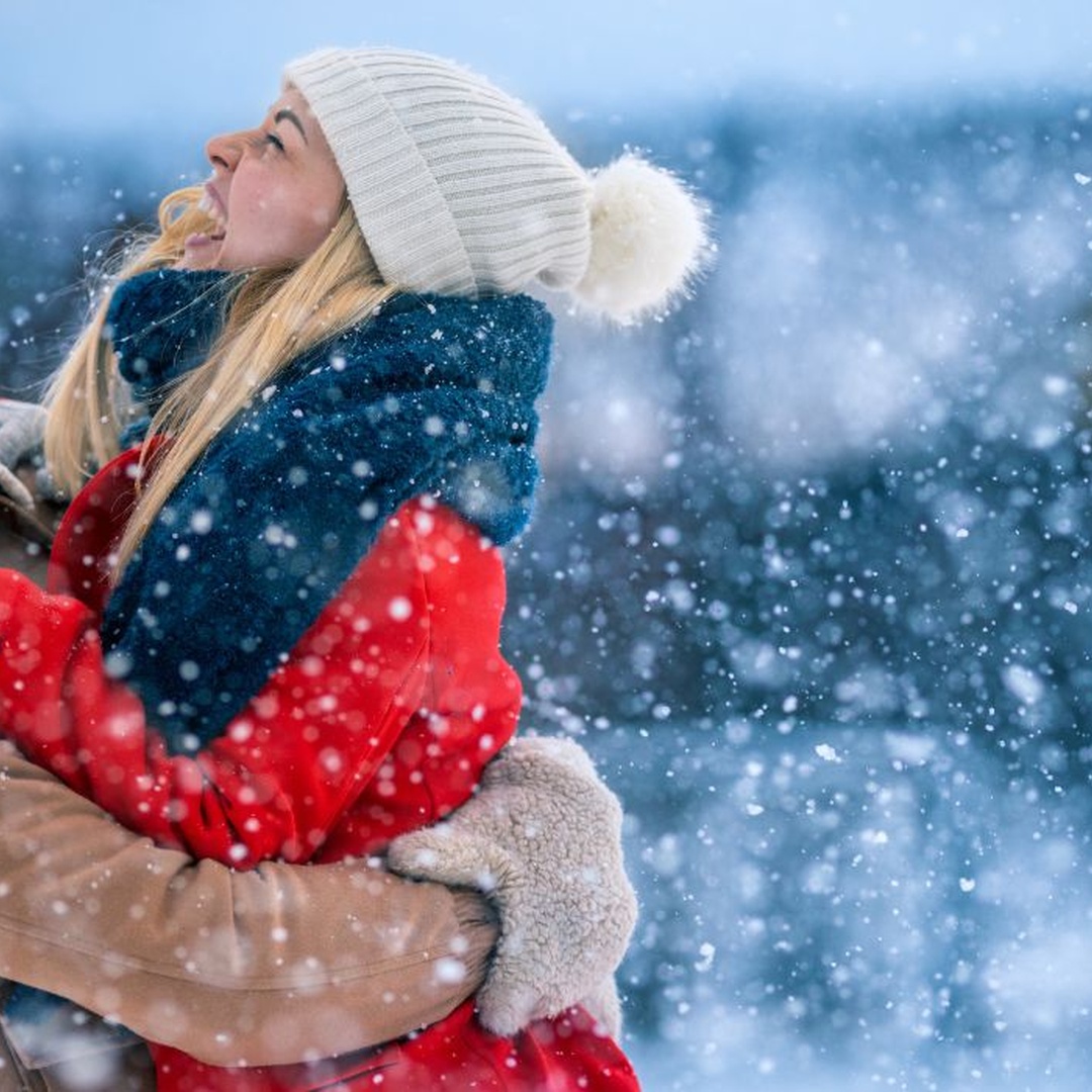 A man wearing a red beanie holding a woman wearing a white beanie in between his arms under the snow.