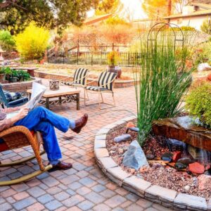 A man in a sweater vest sits in a rocking chair and reads a newspaper in a large, brick-lined patio with a garden.