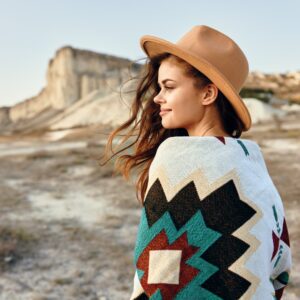 A woman in a tan, wide-brimmed hat and a Southwestern-style sweater stands outdoors, with mountains nearby.