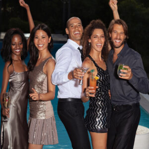 A group of young adults dressed for a party are smiling and laughing together while standing beside a pool.