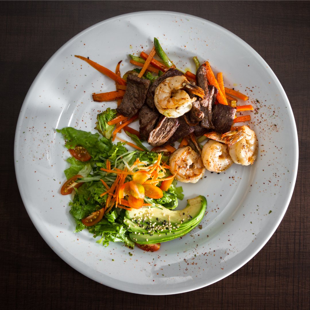 A surf and turf dinner perfectly plated on a white plate. Steak, shrimp, veggies, and a side salad make up the dish.