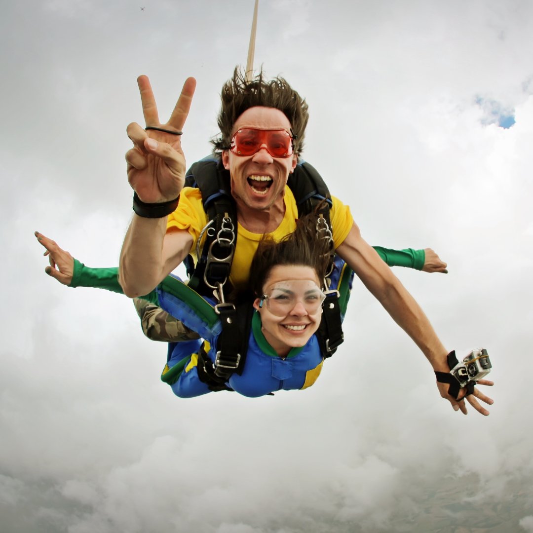 Two people skydiving with one holding up a peace sign. They are freefalling through the sky with dense clouds in the background.