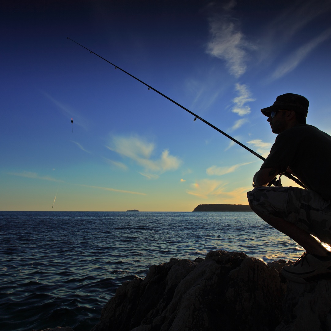 The silhouette of a man crouching on a rock near some water with a fishing pole in hand. The sun sets in the background.