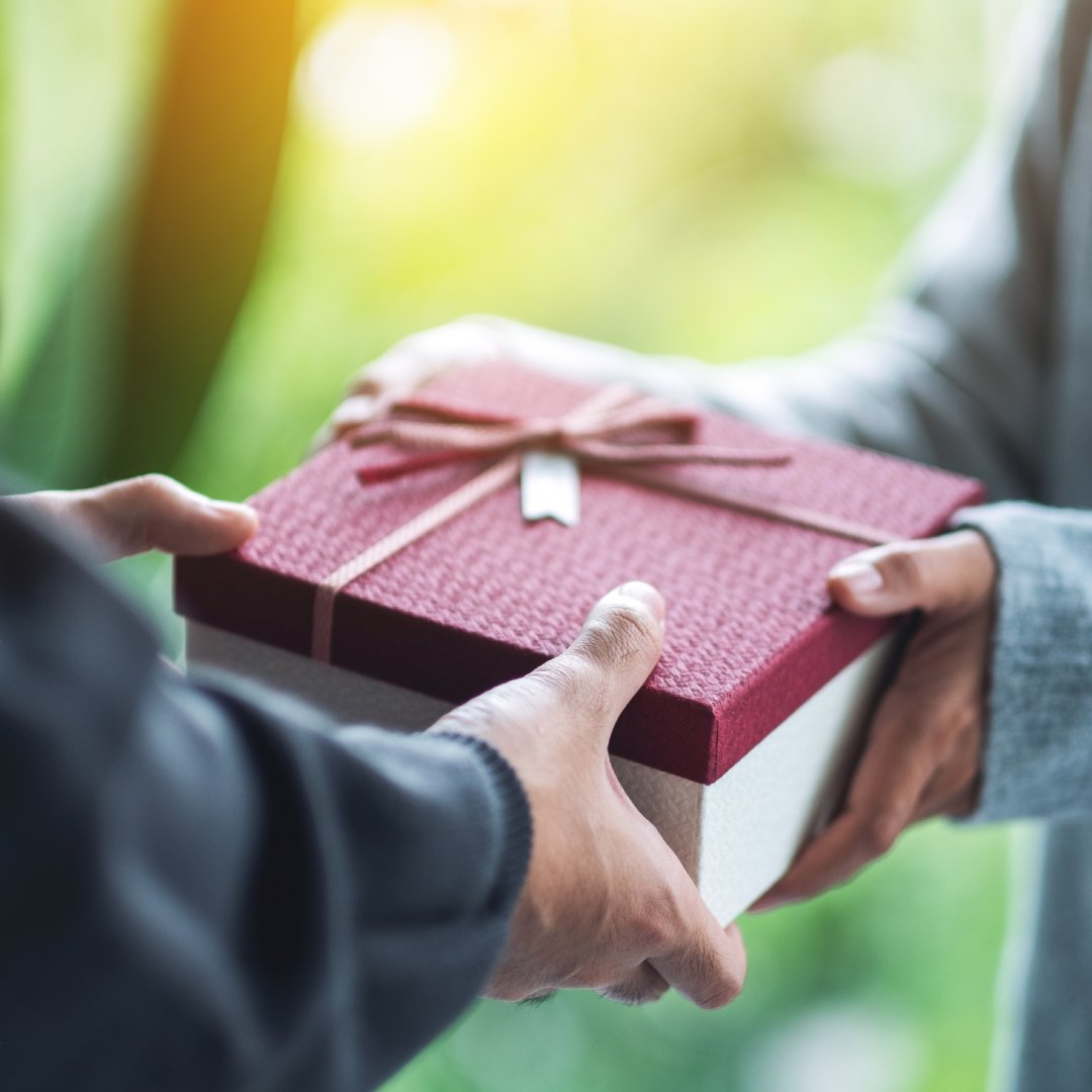 A close-up of two people exchanging a gift. The box is white with a red lid and a perfectly wrapped red ribbon on top.