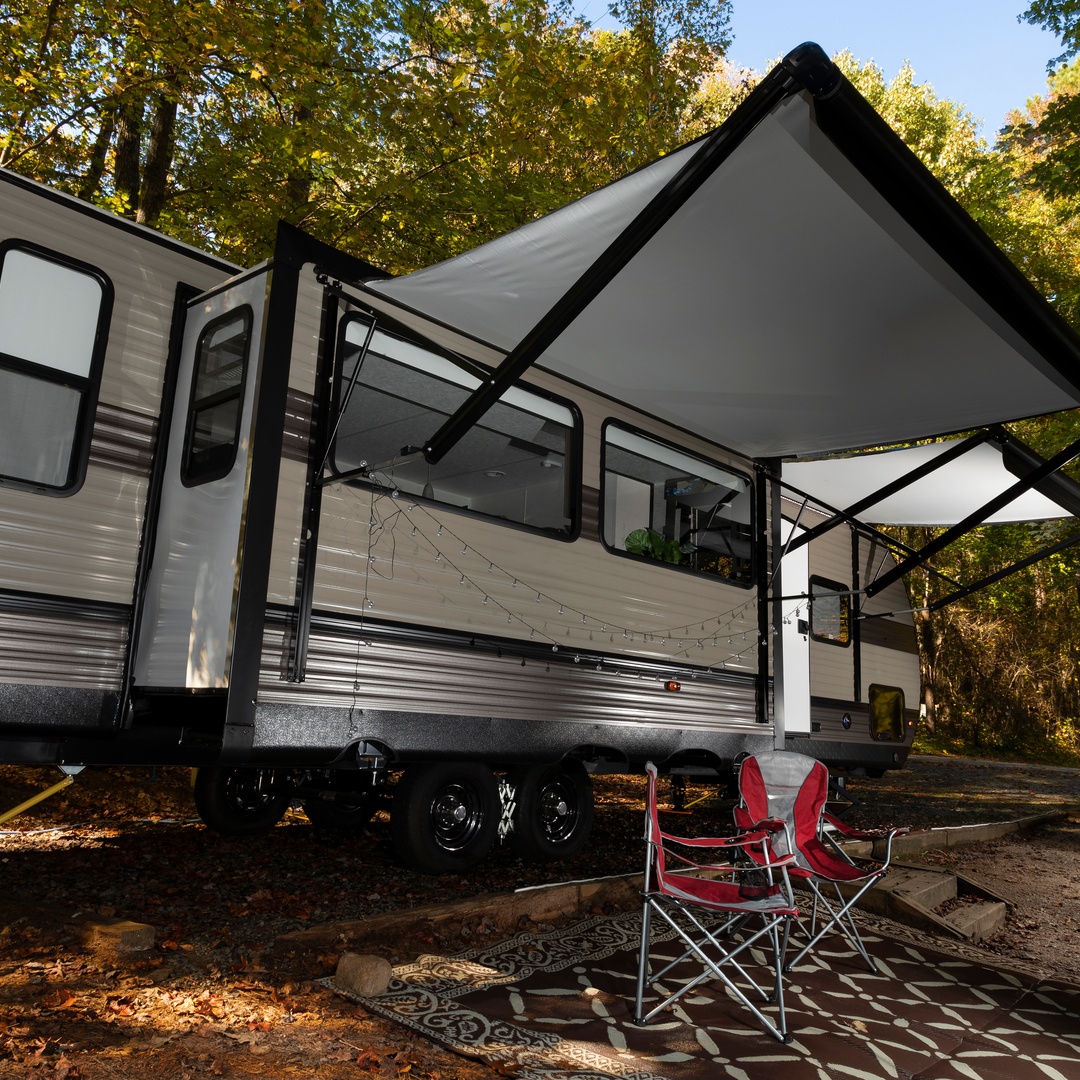 An RV is parked in an area surrounded by green trees with red leaves on the ground. A rug and two chairs are outside the RV.