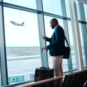 A man looks at his phone as he stands inside of an airport. There is a place taking off right outside the window.