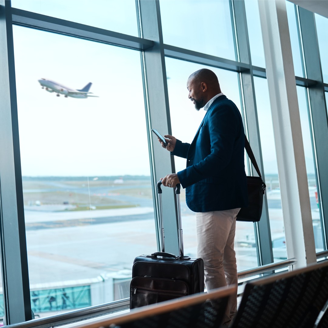 A man looks at his phone as he stands inside of an airport. There is a place taking off right outside the window.