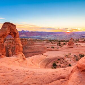 A vibrant sunset frames Delicate Arch in a desert landscape, with red sandstone, distant mountains, and a colorful sky.