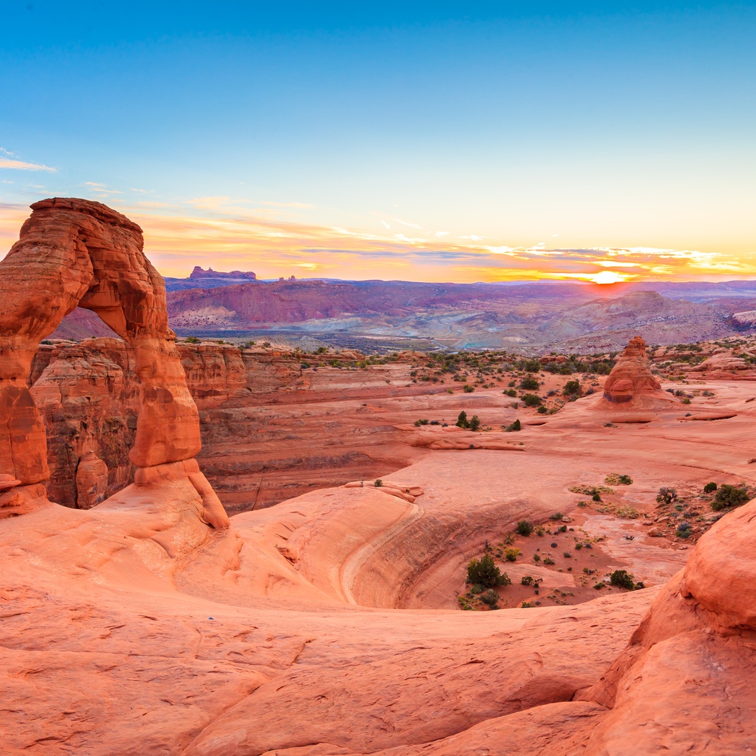 A vibrant sunset frames Delicate Arch in a desert landscape, with red sandstone, distant mountains, and a colorful sky.