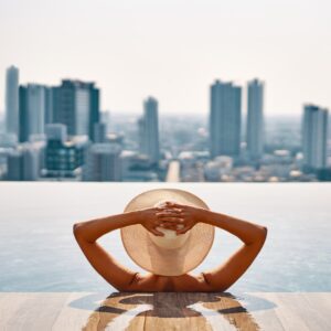 A rear view of a person with a straw hat sitting in a pool as they look out toward the city skyline on a sunny day.