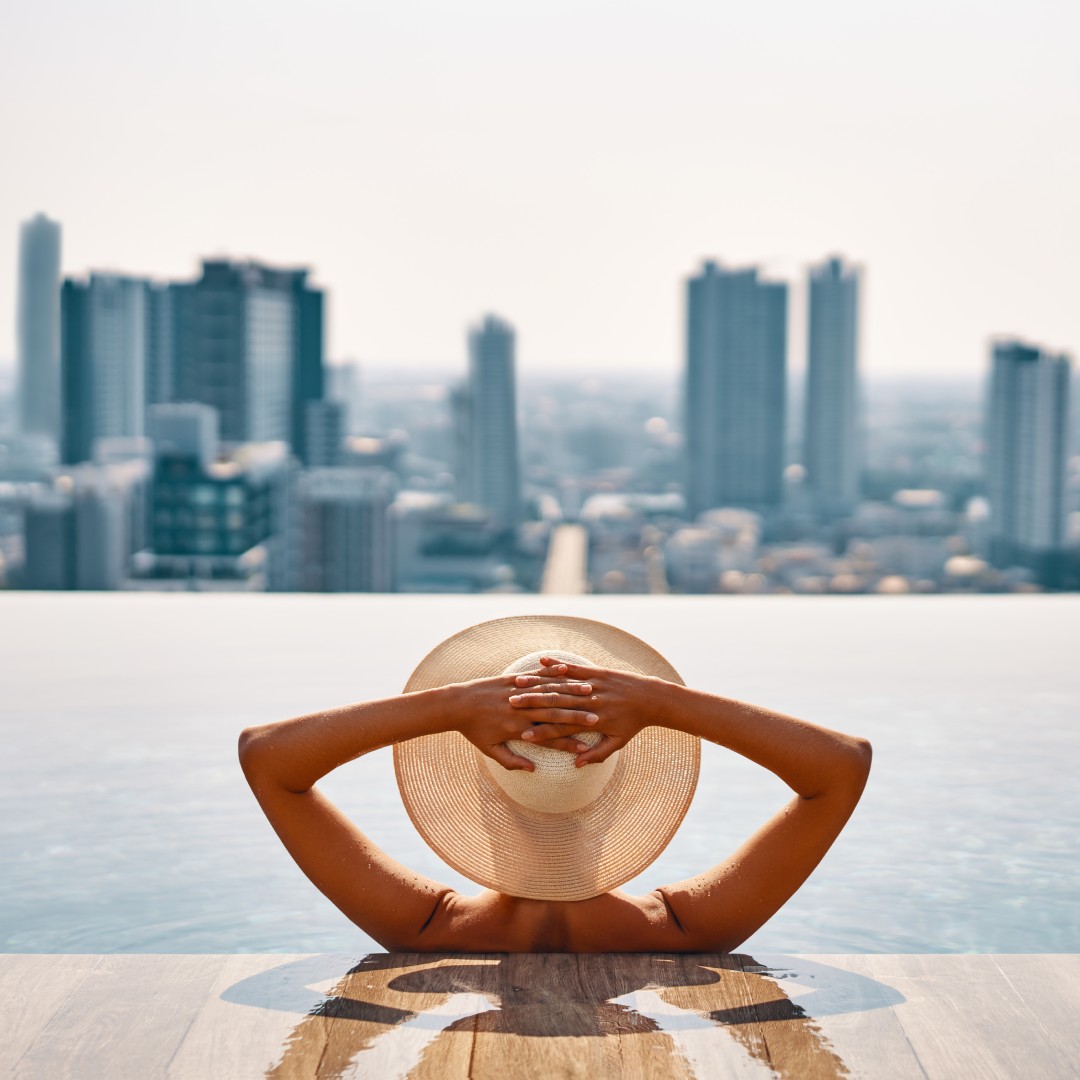 A rear view of a person with a straw hat sitting in a pool as they look out toward the city skyline on a sunny day.