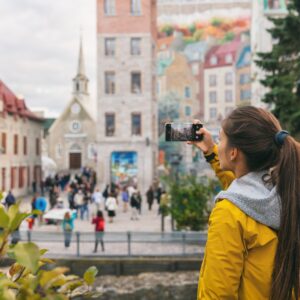A woman wearing a yellow jacket stands in front of a tourist attraction. She takes a photo of a mural with her phone.