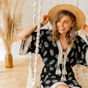 A woman wearing a black and white boho dress and a straw hat smiles while sitting on a white hammock.