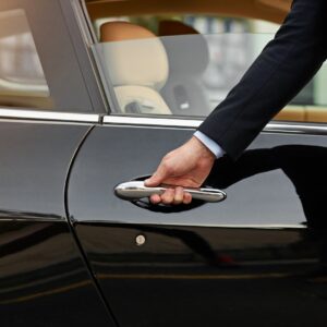 A man in a black suit holds the door handle of a shiny black car, preparing to open the door.