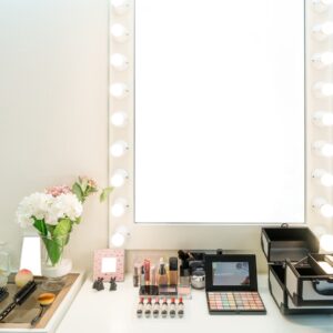 Light bulbs outline the rectangular mirror of a makeup vanity. Eyeshadow, foundation, and makeup brushes are on the surface.