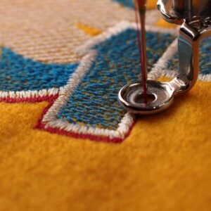 The needle of a sewing machine positioned on top of a piece of fabric. The fabric is orange, white, and blue.