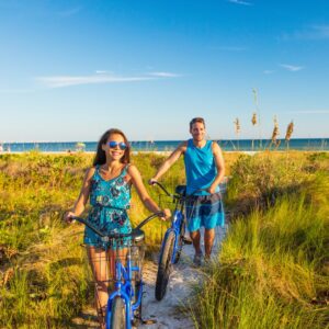 A man and woman walk blue bikes down a sandy path with tall grass on either side of them. The blue ocean is in the background.