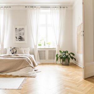 A spacious bedroom with very large windows letting in natural light. The walls and room accents are white with chevron wood flooring.