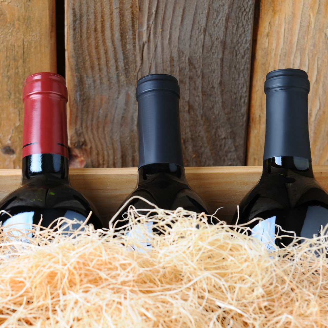 Four wine bottles with red and black caps nestled in straw inside a wooden crate against rustic plank wall