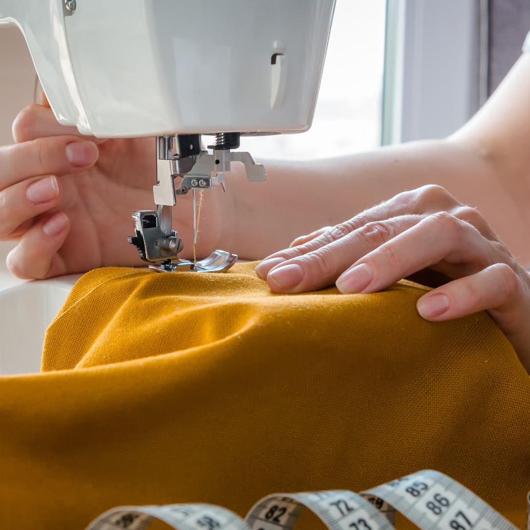 A person guides mustard fabric beneath a sewing machine needle, with a measuring tape and thread resting on the table nearby.