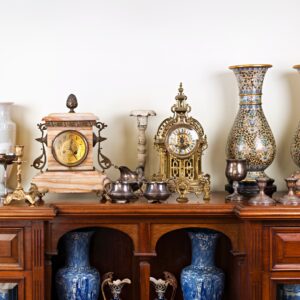 An antique wooden sideboard topped with clocks, patterned vases, silver cups, and decorative ceramics against a cream wall.