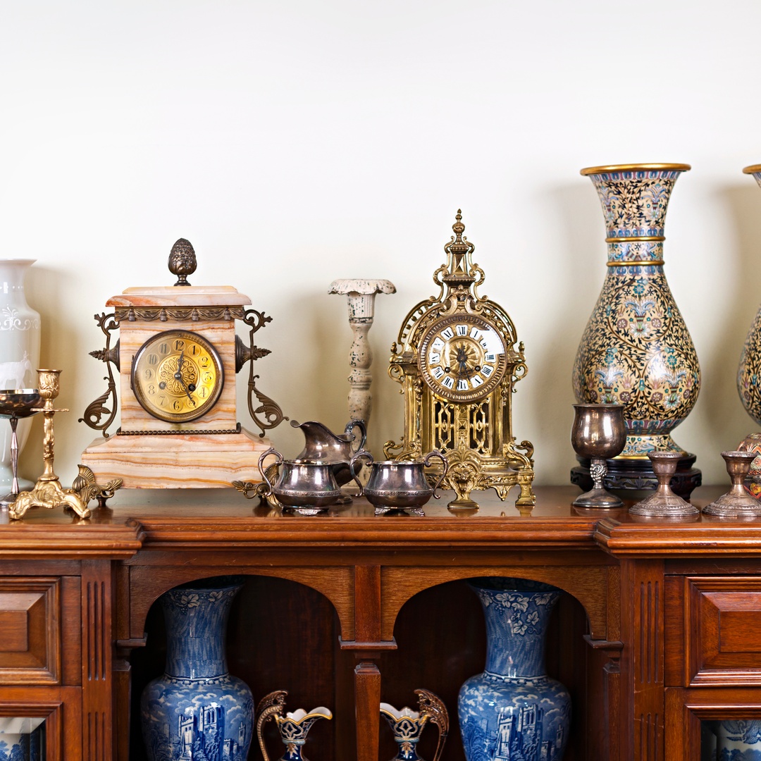 An antique wooden sideboard topped with clocks, patterned vases, silver cups, and decorative ceramics against a cream wall.