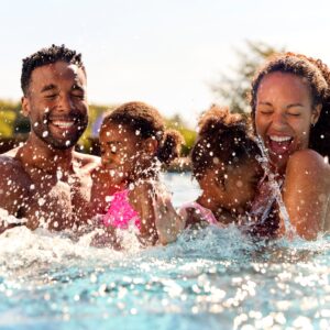 A young Black family on summer vacation, featuring two girls being held by their parents in a swimming pool and splashing.