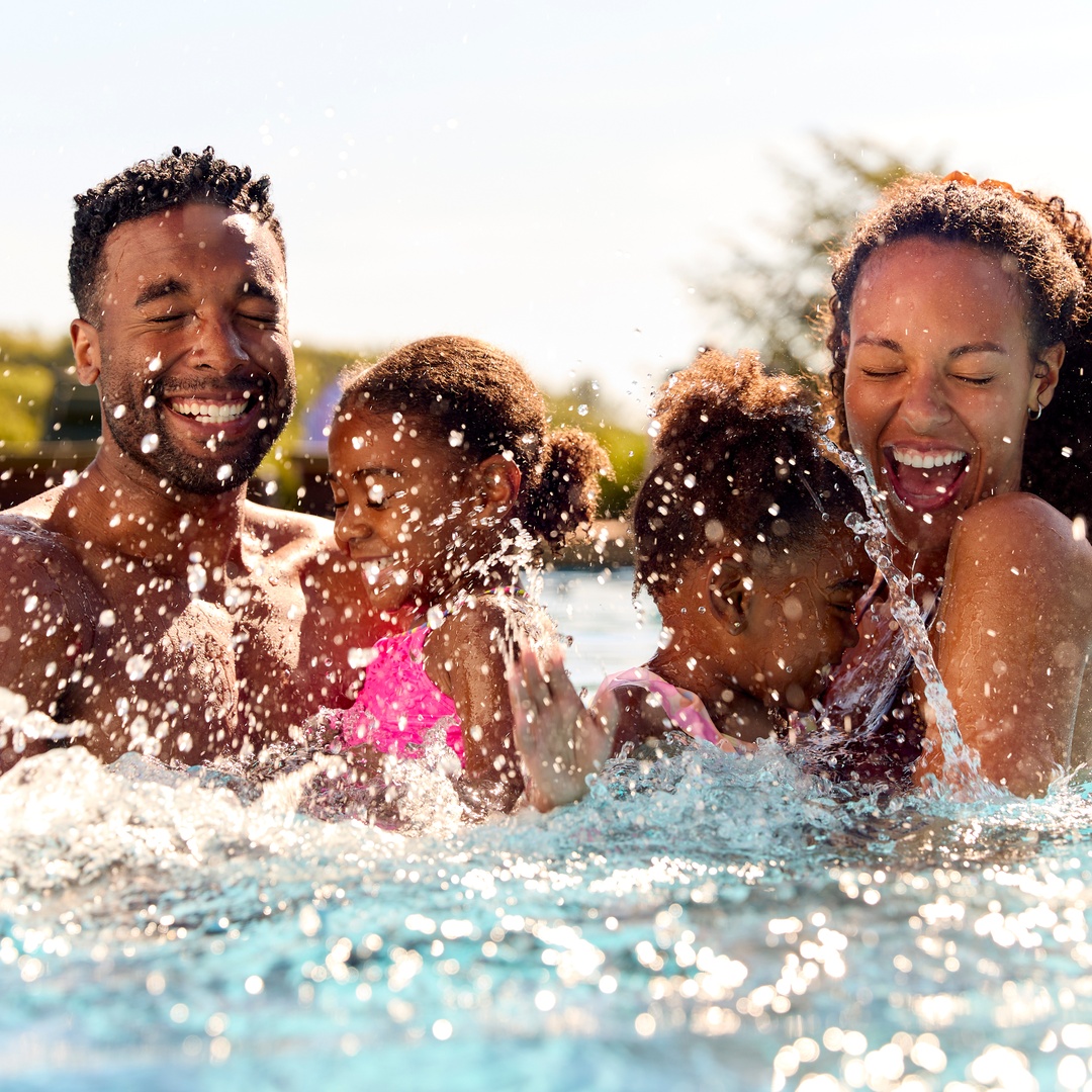 A young Black family on summer vacation, featuring two girls being held by their parents in a swimming pool and splashing.