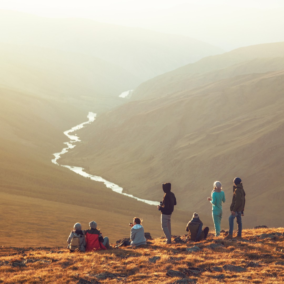 A group of hikers sits and stands on a mountain ridge, looking down at a valley with a river winding below.