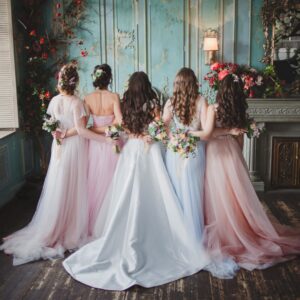 The backsides of five women standing in formal dresses inside a decorated room with flowers and a fireplace.