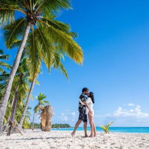 A man and a woman wearing beachy clothing are standing on a beach by palm trees, embracing and kissing.