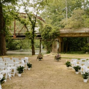 White chairs for wedding guests in front of a small outdoor altar. There is a river to the left and a bridge in the background.